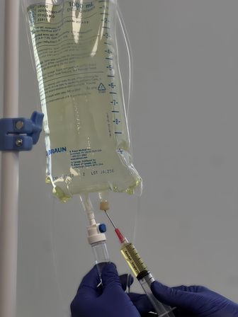 Gloved hands withdraw medication with a syringe from a 1000 mL IV infusion bag hanging on an IV pole in a clinical/hospital setting.