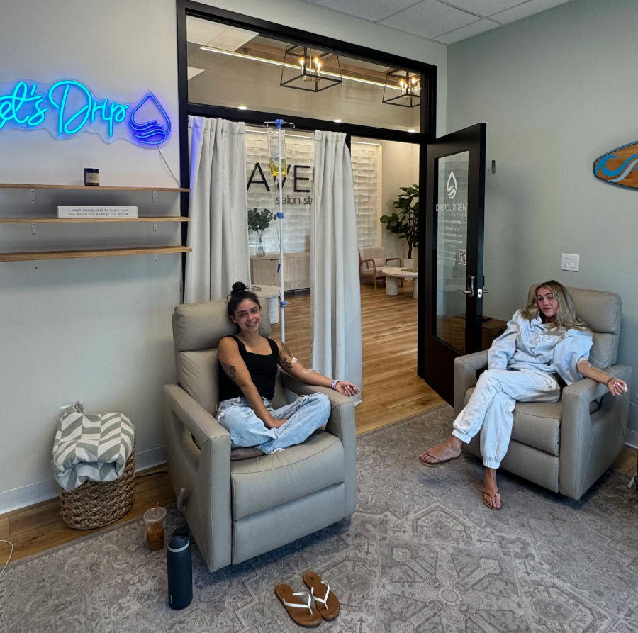 Two people relaxing in beige recliner chairs in a bright wellness lounge receiving IV drip therapy, modern spa interior with neon sign, wooden floors and potted plants.