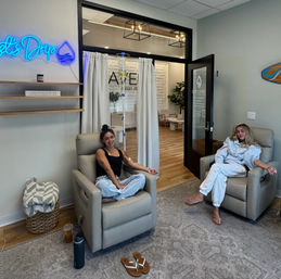 Two people relaxing in beige recliner chairs in a bright wellness lounge receiving IV drip therapy, modern spa interior with neon sign, wooden floors and potted plants.