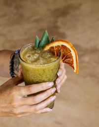 Refreshing green tropical smoothie in a glass, held by hands and garnished with pineapple leaves and a dried orange slice against a warm beige backdrop