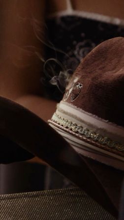 Close-up of a brown suede cowboy hat with braided gold-trim ribbon and embroidered detail, wisps of smoke curling above the brim in warm, moody lighting.