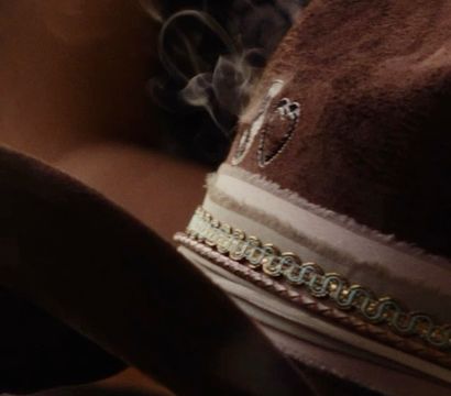 Close-up of a brown suede cowboy hat with braided gold-trim ribbon and embroidered detail, wisps of smoke curling above the brim in warm, moody lighting.