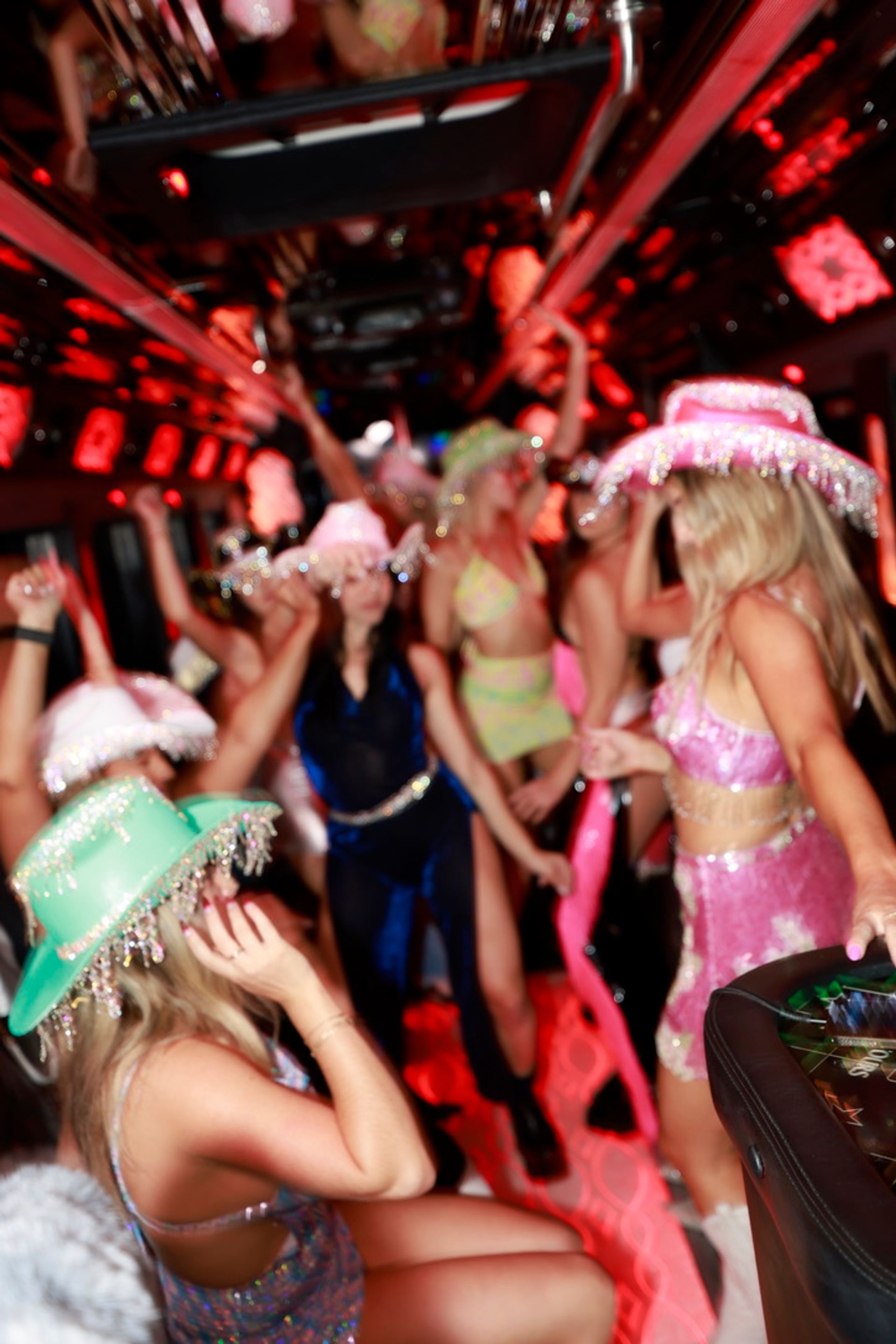 Crowded party bus interior with people dancing under red neon lights, many wearing sparkly cowboy hats and glittery crop-top outfits in a motion-blurred, energetic celebration.