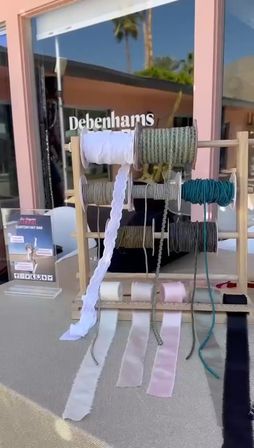 Outdoor craft pop-up with a wooden rack of ribbon spools and cords — white lace, sage and teal ropes and pink and black satin strips draping over a table in front of a storefront reflecting palm trees.