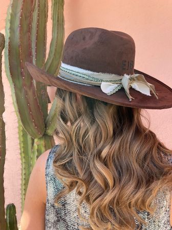 Back view of a person with long wavy blonde hair wearing a brown wide‑brim boho hat with ribbon trim, standing by a tall cactus against a peach stucco wall for a southwestern desert vibe.