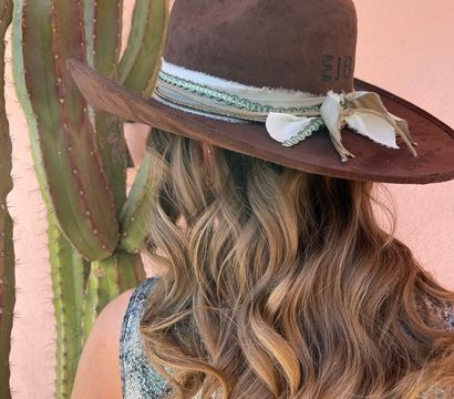 Back view of a person with long wavy blonde hair wearing a brown wide‑brim boho hat with ribbon trim, standing by a tall cactus against a peach stucco wall for a southwestern desert vibe.