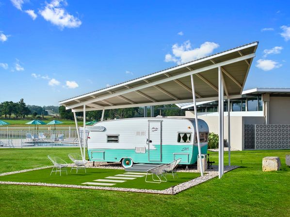 Retro turquoise-and-white travel trailer parked under a modern metal canopy on a manicured lawn with outdoor seating, poolside umbrellas and bright blue sky.