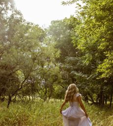 Sunlit woodland meadow with a blonde woman in a flowing pastel dress walking through tall grass, surrounded by green trees — pastoral countryside nature scene.