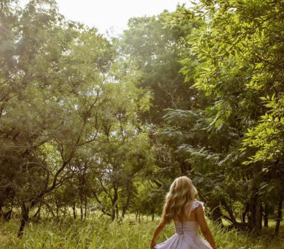 Sunlit woodland meadow with a blonde woman in a flowing pastel dress walking through tall grass, surrounded by green trees — pastoral countryside nature scene.
