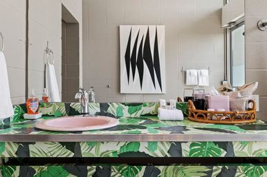 Tropical leaf-print bathroom vanity with pink sink and chrome faucet, wicker tray of toiletries, rolled towel, mirror and abstract black-and-white wall art