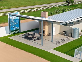 Sunlit modern covered patio with stone fireplace and black lounge seating under a steel pergola beside a metal-roofed outbuilding, corrugated water tank, decorative mural, manicured green lawn and white fence along a country road.