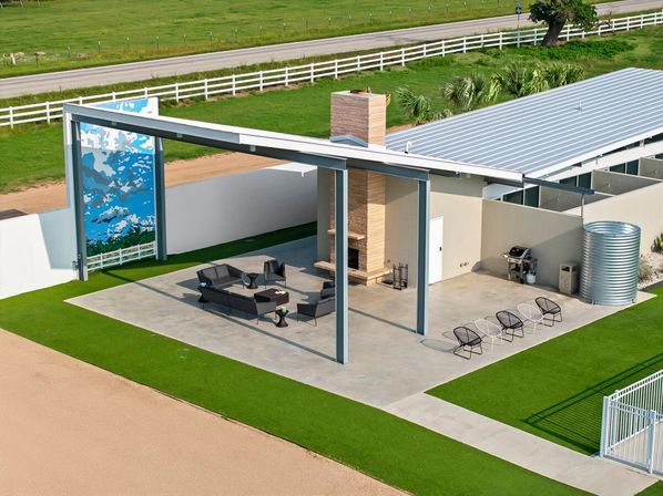 Sunlit modern covered patio with stone fireplace and black lounge seating under a steel pergola beside a metal-roofed outbuilding, corrugated water tank, decorative mural, manicured green lawn and white fence along a country road.