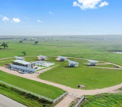 Aerial view of a rural property with modern tiny cabins arranged around a circular dirt road encircling a central white pavilion and pool, set on green pasture with white fencing, a country road and expansive farmland under a bright blue sky.