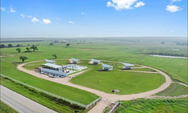 Aerial view of a rural property with modern tiny cabins arranged around a circular dirt road encircling a central white pavilion and pool, set on green pasture with white fencing, a country road and expansive farmland under a bright blue sky.