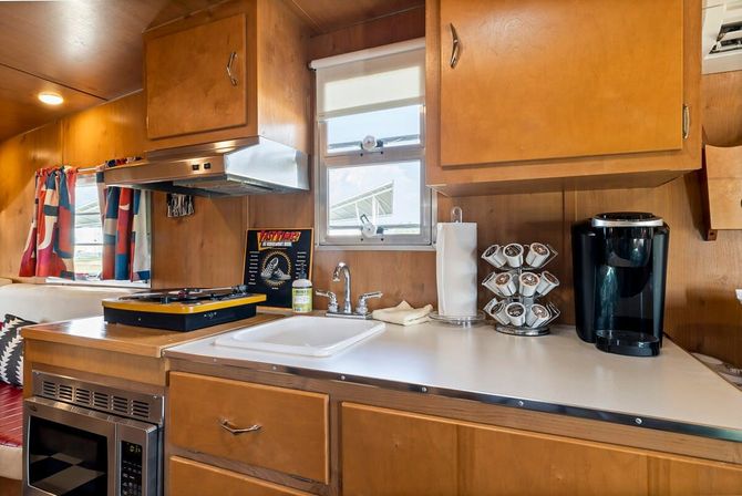 Compact retro camper kitchenette with warm wood cabinets, small sink and faucet, two-burner stove under a range hood, microwave drawer, black single-serve coffee maker with K-cup carousel, paper towel holder and colorful curtains by the window.