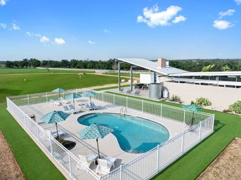 Fenced outdoor swimming pool with turquoise umbrellas and white lounge chairs on a concrete deck, a modern metal-roof pavilion nearby, and rolling green fields under a bright blue sky — inviting rural leisure scene.
