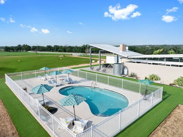 Fenced outdoor swimming pool with turquoise umbrellas and white lounge chairs on a concrete deck, a modern metal-roof pavilion nearby, and rolling green fields under a bright blue sky — inviting rural leisure scene.