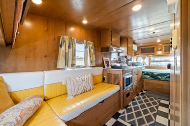 Cozy retro camper RV interior with yellow vinyl bench seating, wood-paneled walls, compact kitchenette with microwave, rear bed, patterned curtains and black-and-white checkered floor