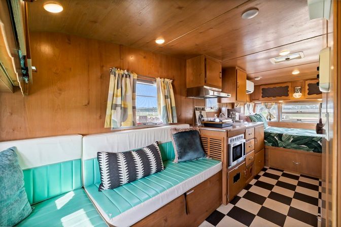Bright retro camper interior with wood-paneled walls, teal vinyl bench seating, black-and-white checkerboard floor, compact kitchenette and a cozy bed by waterfront windows