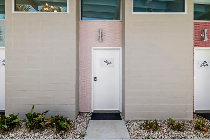 Retro mid-century motel entrance with white door set in a pink-tiled inset, beige cinderblock facade, silver wall sconce above, pebbled landscaping and low tropical plants