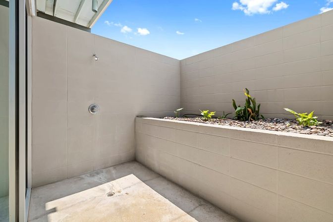Private sunny courtyard with a wall-mounted outdoor shower, concrete floor and drain, raised pebble-filled planter with green plants and high beige block walls under a bright blue sky.