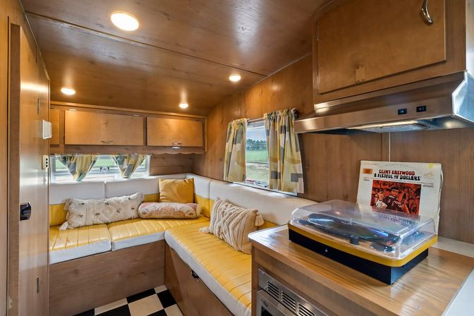 Cozy retro travel trailer interior with yellow vinyl L-shaped bench and throw pillows, wood-paneled walls and overhead cabinets, patterned curtains, black-and-white checkered floor, and a turntable on the countertop.