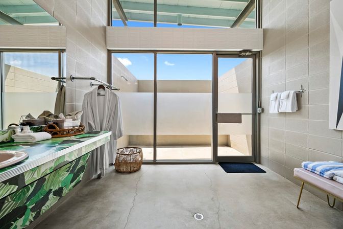 Sunny modern spa changing room with frosted glass sliding door to a private outdoor shower patio, hanging white robes, wicker basket, tropical-leaf vanity, folded towels on a bench and concrete floor