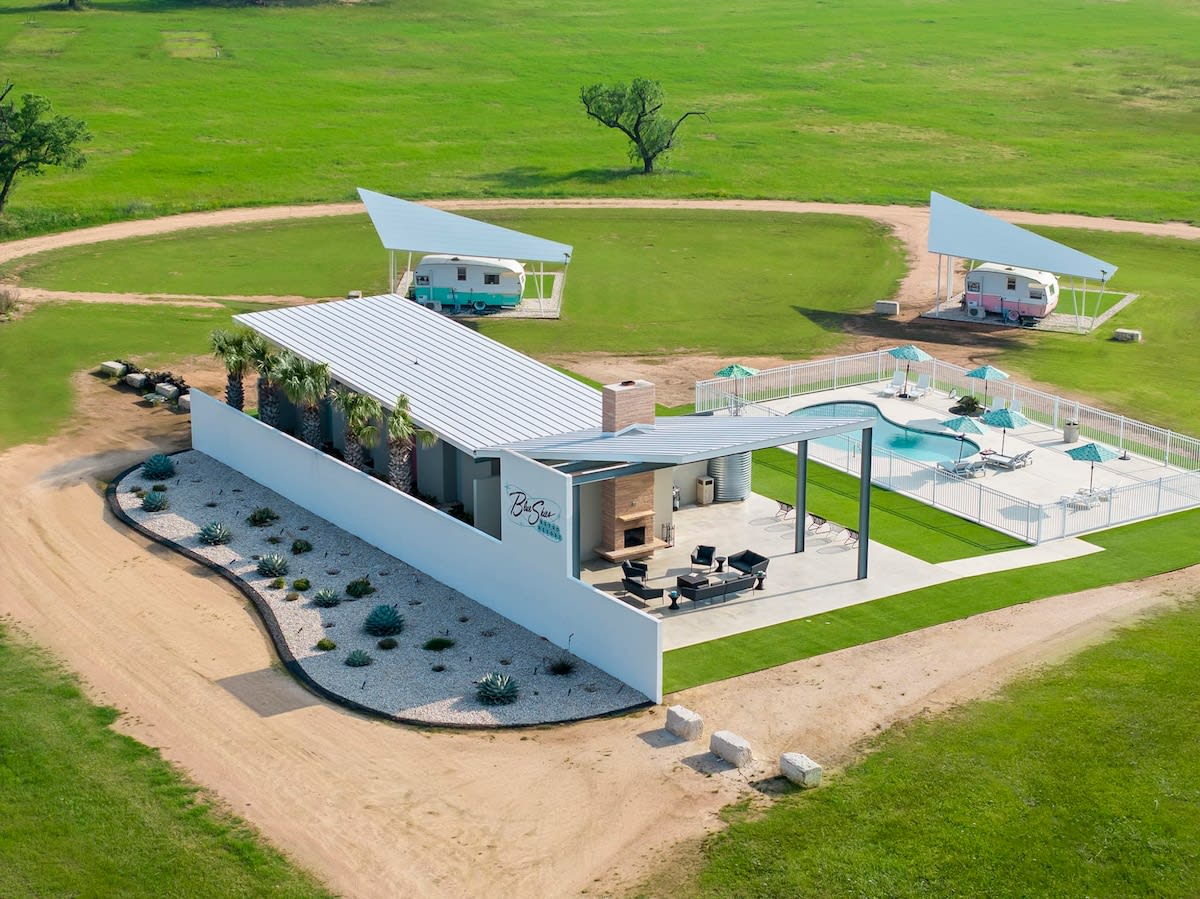 Aerial view of a sunny countryside glamping site with a modern pool and patio featuring an outdoor fireplace, desert rock landscaping, and two pastel vintage campers parked under triangular shade canopies on a wide green lawn.