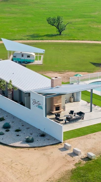Aerial view of a sunny countryside glamping site with a modern pool and patio featuring an outdoor fireplace, desert rock landscaping, and two pastel vintage campers parked under triangular shade canopies on a wide green lawn.