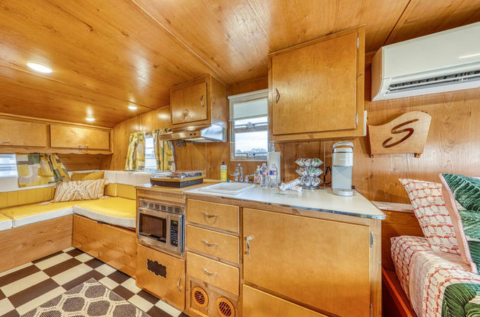 Cozy retro wood-paneled travel trailer interior with compact kitchenette — sink, microwave, coffee maker, overhead cabinets, yellow L-shaped cushioned seating, air conditioner, and black-and-white checkered floor.