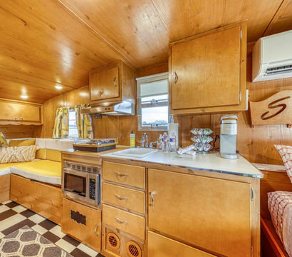 Cozy retro wood-paneled travel trailer interior with compact kitchenette — sink, microwave, coffee maker, overhead cabinets, yellow L-shaped cushioned seating, air conditioner, and black-and-white checkered floor.