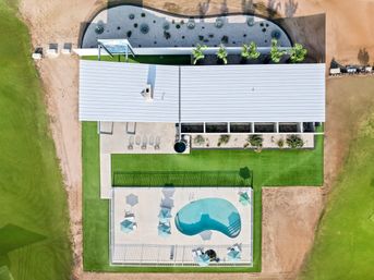 Aerial drone view of a modern single-story building with metal roof, manicured lawn, desert-style planters and a fenced kidney-shaped pool with umbrellas and lounge chairs.
