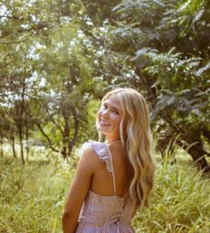 Smiling blonde woman in a pastel sundress looks over her shoulder in a sunlit meadow surrounded by trees — outdoor summer golden-hour portrait