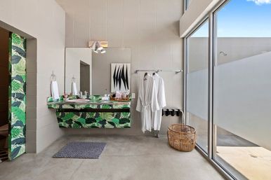 Sunlit modern bathroom with a leafy green patterned floating vanity and large mirror, concrete floor, floor-to-ceiling glass wall with frosted panel, two white bathrobes on a rail, wicker laundry basket and gray bath mat.