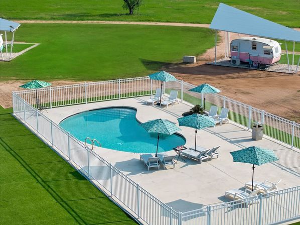 Aerial view of a fenced kidney-shaped outdoor pool with turquoise umbrellas and white lounge chairs on a sunny concrete deck, set amid bright green lawn with a retro pink camper parked under a covered shelter in a rural setting.