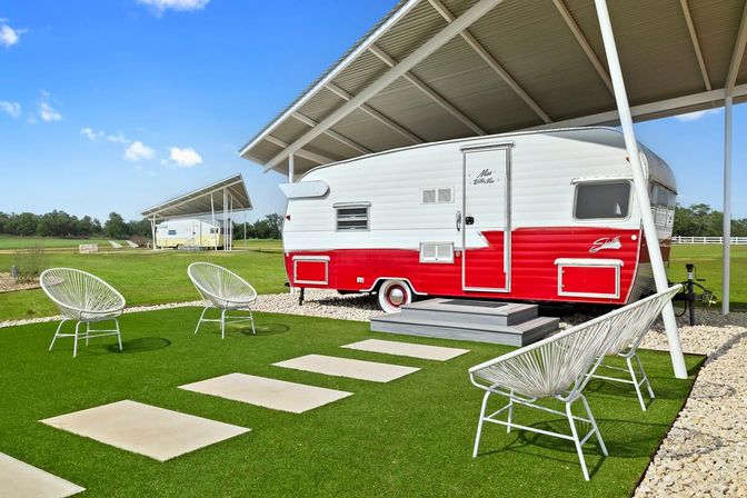 Retro red-and-white travel trailer under a metal canopy on a manicured green lawn with white lounge chairs, stepping stones, and blue sky — covered glamping RV site.