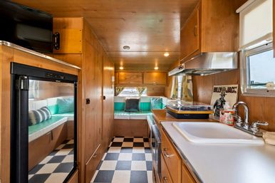 Bright vintage camper interior with wood-paneled walls, retro black-and-white checkerboard floor, compact kitchenette with sink and range, teal vinyl bench seating and cozy dinette at the rear.