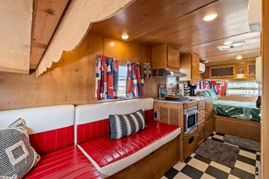 Cozy retro camper interior with wood-paneled walls, red-and-white vinyl bench, patterned pillows, black-and-white checkered floor, compact kitchenette with microwave and range hood, colorful curtains, and a green botanical-pattern bed at the rear.