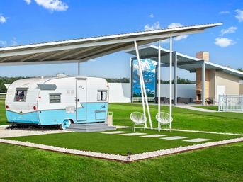 Teal-and-white vintage travel trailer on a manicured green lawn under a modern metal canopy, with two white patio chairs, stepping stones and a covered outdoor lounge area beneath a bright blue sky