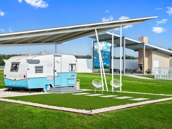 Teal-and-white vintage travel trailer on a manicured green lawn under a modern metal canopy, with two white patio chairs, stepping stones and a covered outdoor lounge area beneath a bright blue sky