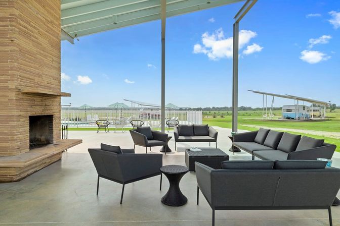 Modern covered patio with black lounge seating and stone fireplace overlooking a pool, open grassy countryside and parked RVs under canopies beneath a bright blue sky.