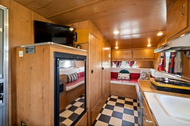 Cozy retro travel trailer interior with wood paneling, checkerboard floor, compact kitchenette, red vinyl dinette and wall-mounted TV.