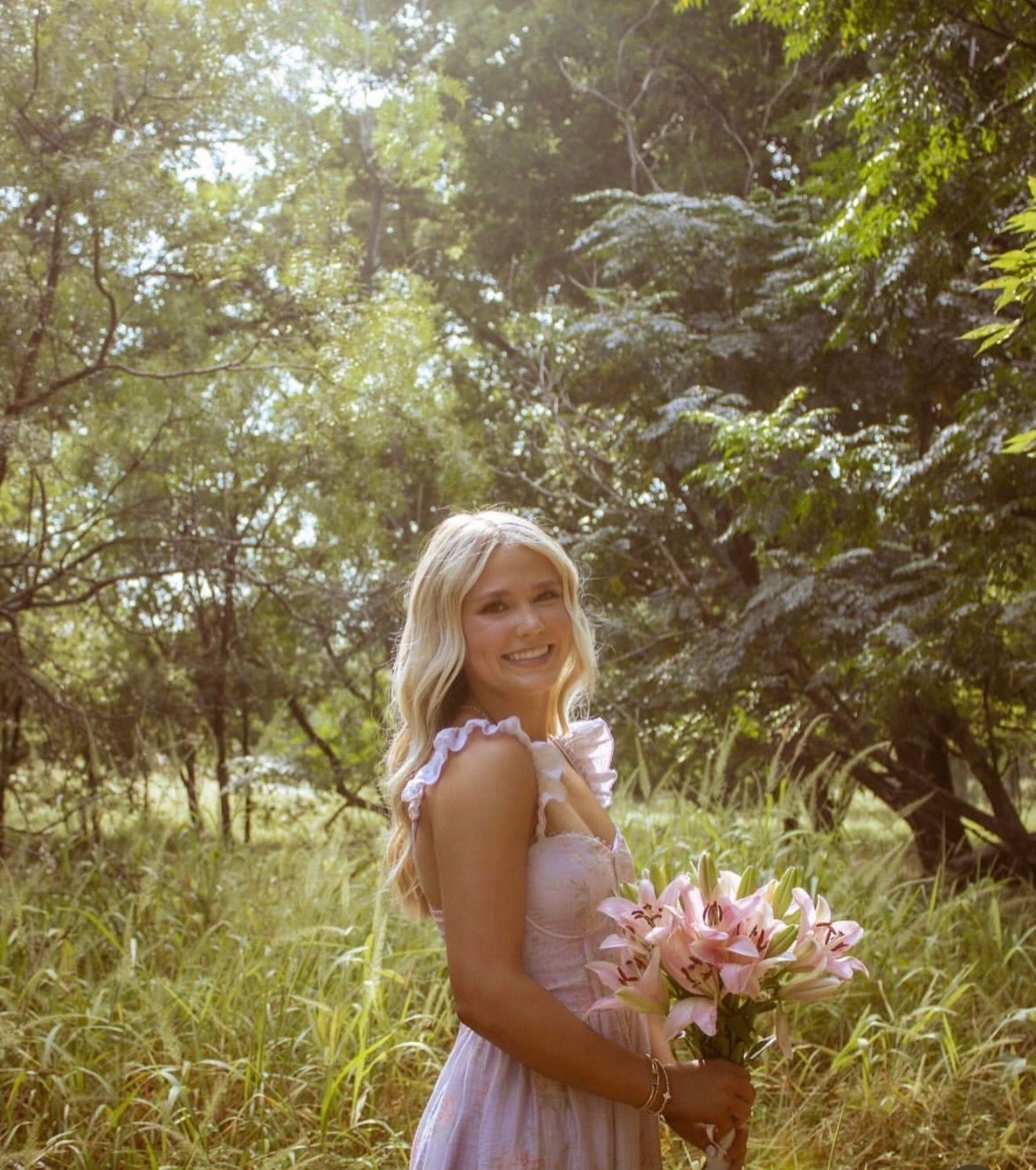 Sunlit outdoor portrait of a smiling woman in a pink summer dress holding a bouquet of pink lilies in a grassy, wooded meadow