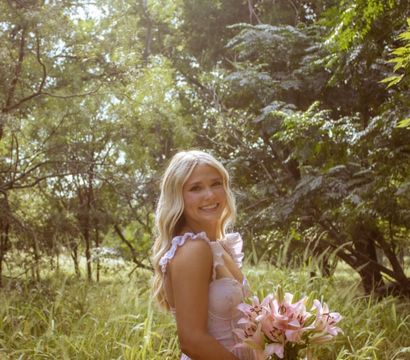 Sunlit outdoor portrait of a smiling woman in a pink summer dress holding a bouquet of pink lilies in a grassy, wooded meadow