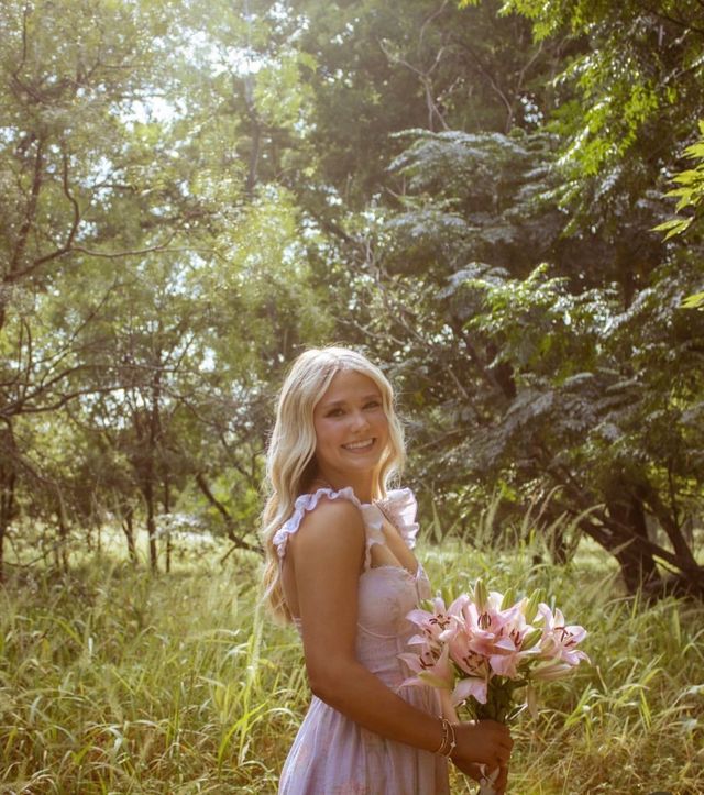 Sunlit outdoor portrait of a smiling woman in a pink summer dress holding a bouquet of pink lilies in a grassy, wooded meadow