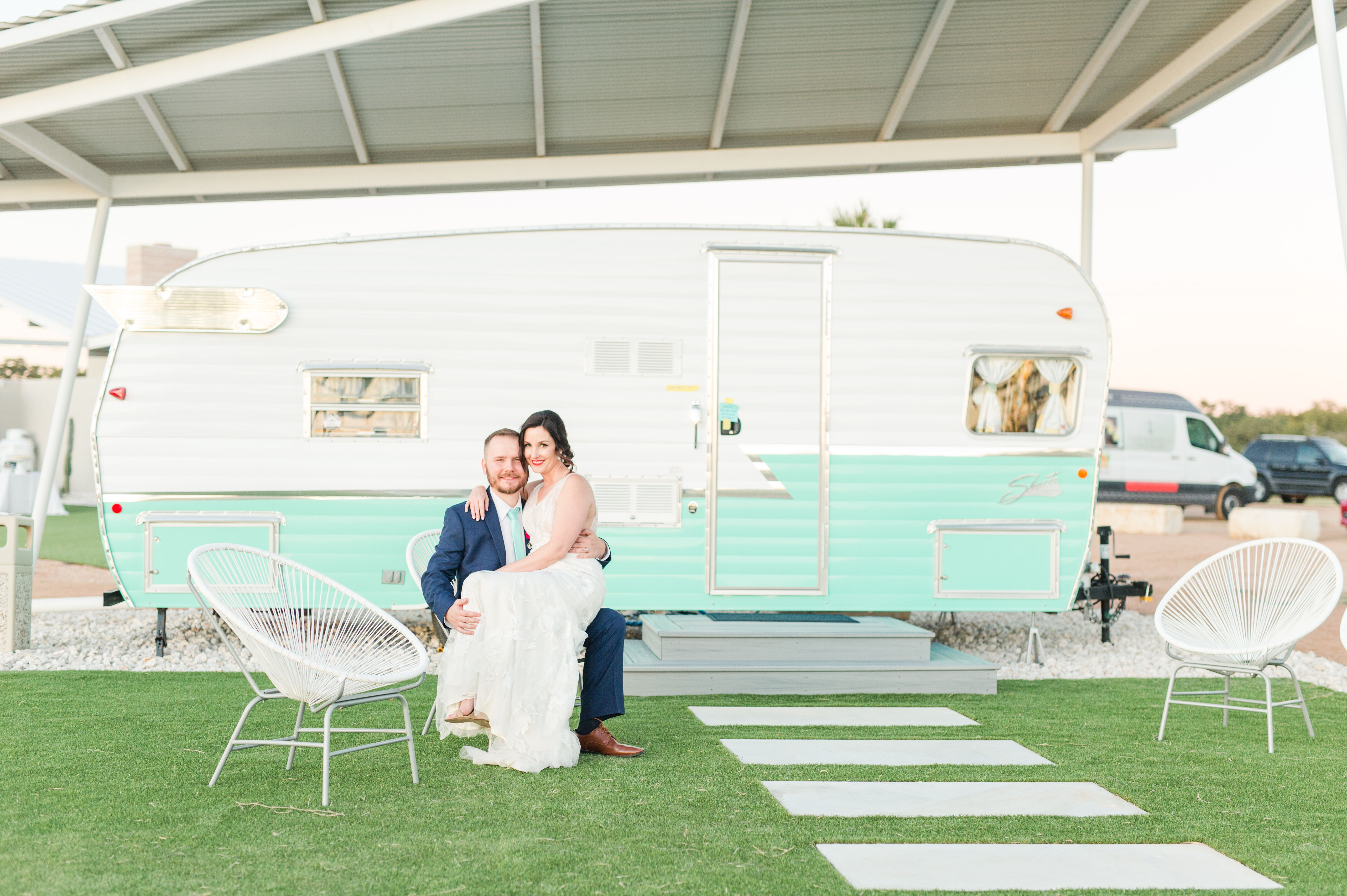 Playful wedding portrait of a bride in a white gown and groom in a navy suit seated on a lawn in front of a mint green vintage camper under a covered carport, with modern patio chairs and stepping stones.
