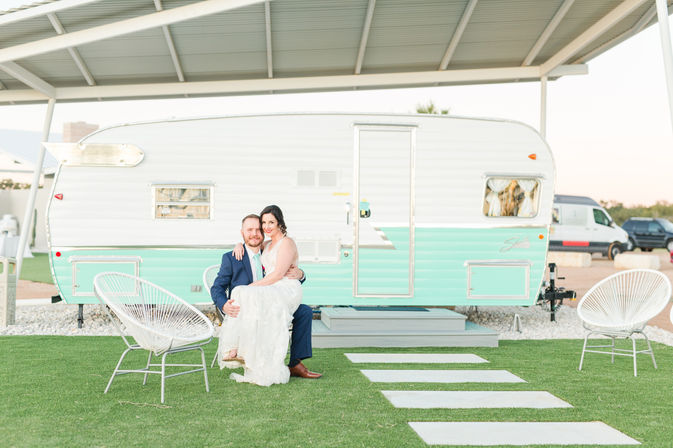 Playful wedding portrait of a bride in a white gown and groom in a navy suit seated on a lawn in front of a mint green vintage camper under a covered carport, with modern patio chairs and stepping stones.