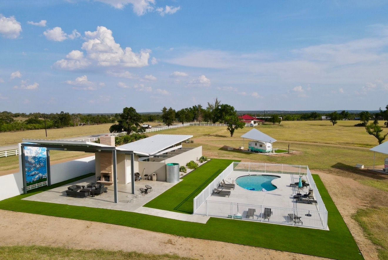 Aerial view of a sunny rural property with a fenced kidney-shaped pool and lounge chairs, a modern covered patio with seating and fireplace under a metal canopy, neat green turf strips, small outbuildings, open grassy fields and a blue sky with scattered clouds.