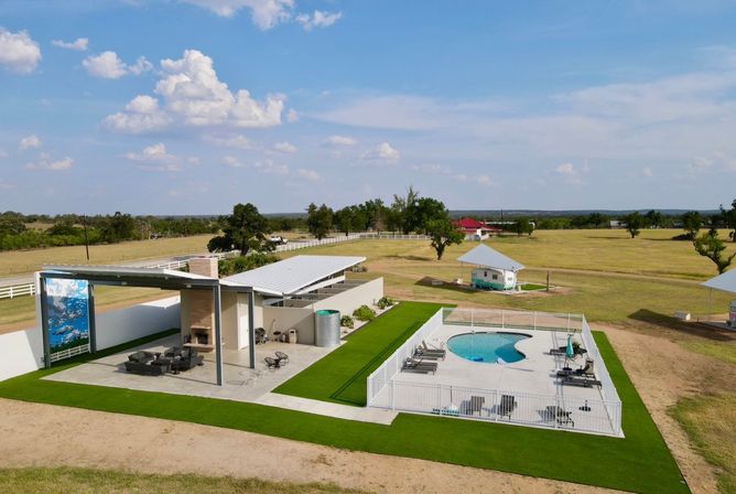 Aerial view of a sunny rural property with a fenced kidney-shaped pool and lounge chairs, a modern covered patio with seating and fireplace under a metal canopy, neat green turf strips, small outbuildings, open grassy fields and a blue sky with scattered clouds.