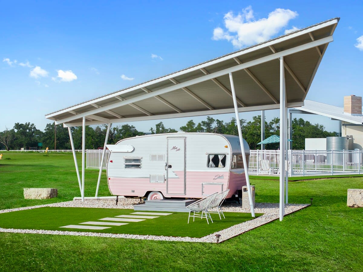 Retro pink-and-white travel trailer parked under a modern metal canopy on a manicured green lawn with stepping stones, white gravel border, two outdoor chairs and a bright blue sky.